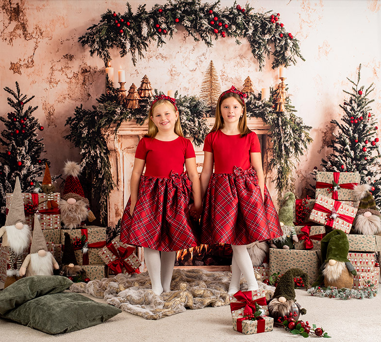 Dve deklici v ujemajočih rdeče-kariranih oblekah stojita pred praznično okrašenim kaminom in se držita za roke; okoli so darila, smrečice in božični palčki.