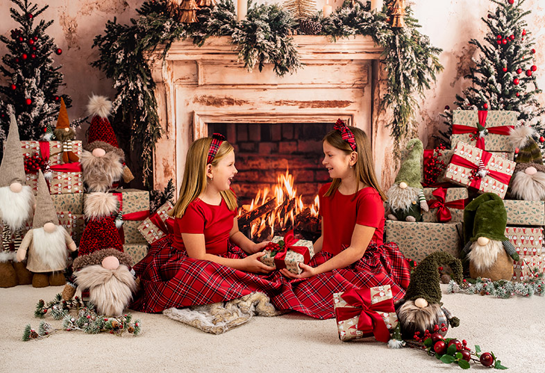 Dve deklici v rdečih kariranih oblekah sedita pred kaminom in si podarjata darila; okoli sta darilne škatle, božični palčki in okrašene smrečice.