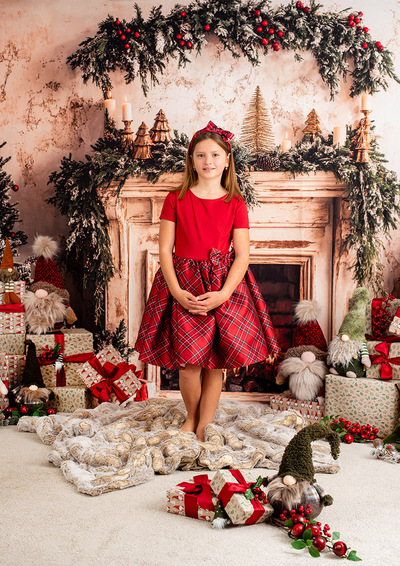 Deklica v rdeči karirani obleki stoji pred prazničnim kaminom, obkrožena z darili in božičnimi palčki.