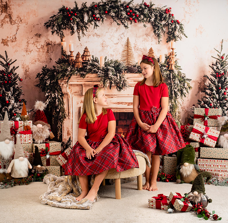 Dve deklici v rdečih kariranih oblekah; ena sedi na tabureju, druga stoji ob njej. Za njima je praznično okrašen kamin z darili, smrečicami in božičnimi palčki.